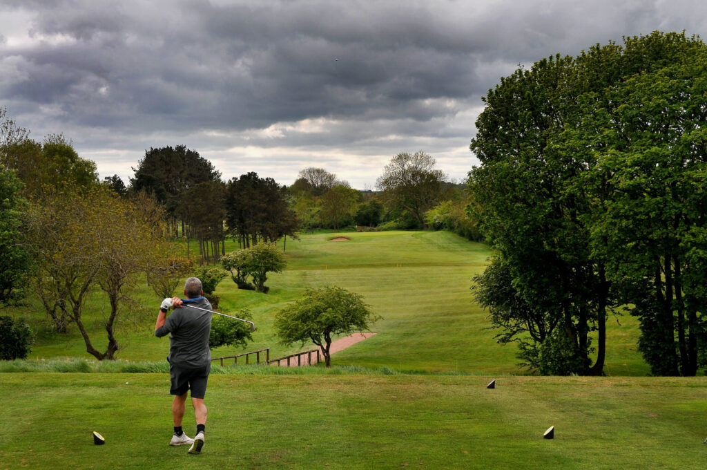 Golfer teeing off with wide view of tree-lined fairway under cloudy skies